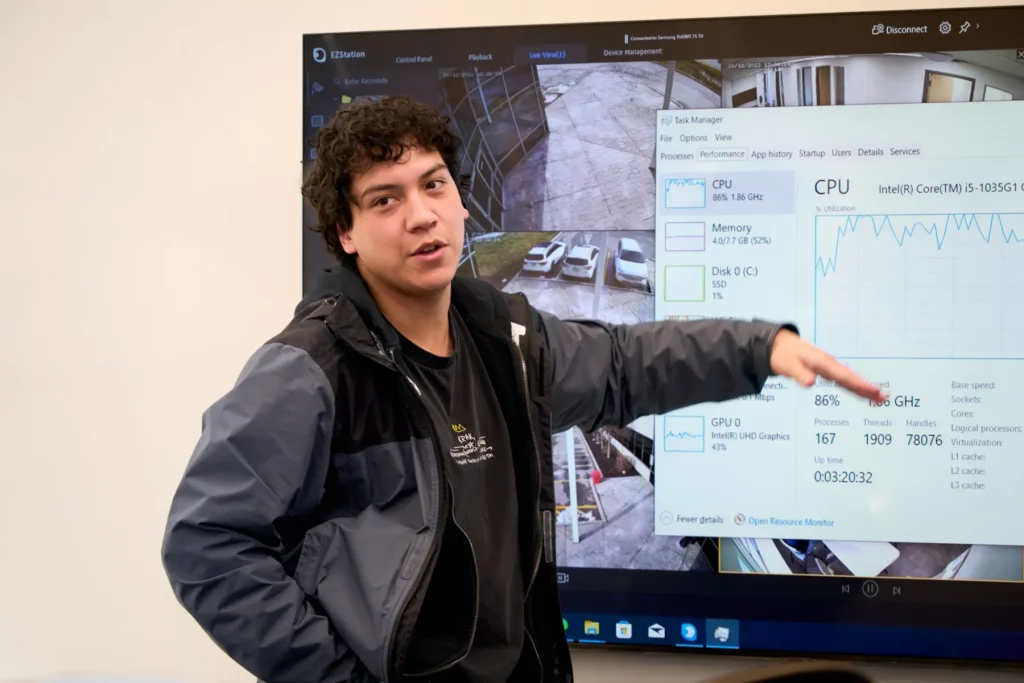 A man in a grey jacket standing in front of a large computer monitor with EZStation and Task Manager opened in the background
